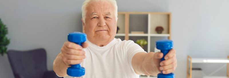 Old man trying to stay active by exercising with blue weights and wearing a white t-shirt